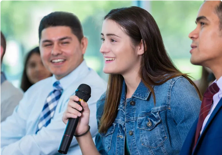 Woman holding a mic and addressing a group of people at a corporate event