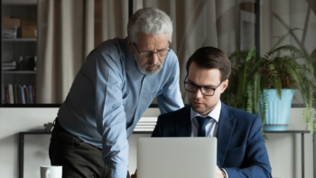 older man standing behind younger man looking at a laptop screen.