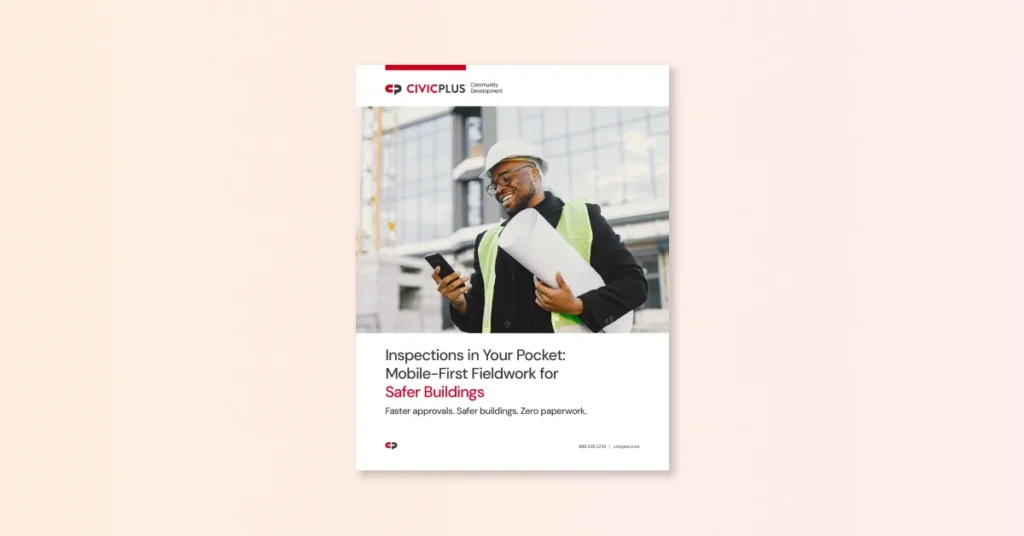 man wearing a construction safety vest and hat holding a roll of paper while smiling, looking at a cell phone in his hand.