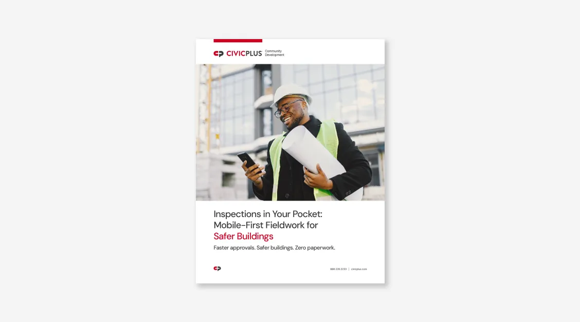 man wearing a construction safety vest and hat holding a roll of paper while smiling, looking at a cell phone in his hand.