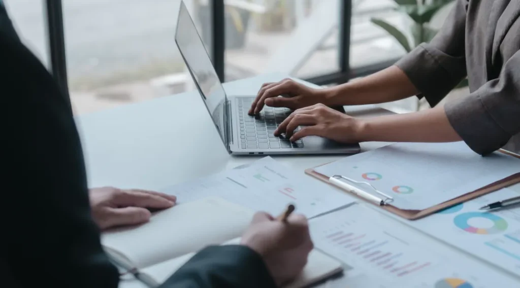 hands at a laptop and a book on a desk.