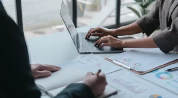 hands at a laptop and a book on a desk.